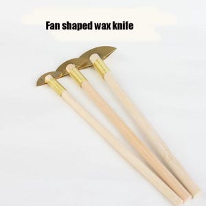 May include: Fan-shaped wax knives with wooden handles and brass-coloured blades. The text "Fan shaped wax knife" is displayed above the knives. The tools are designed for wax carving and sculpting.