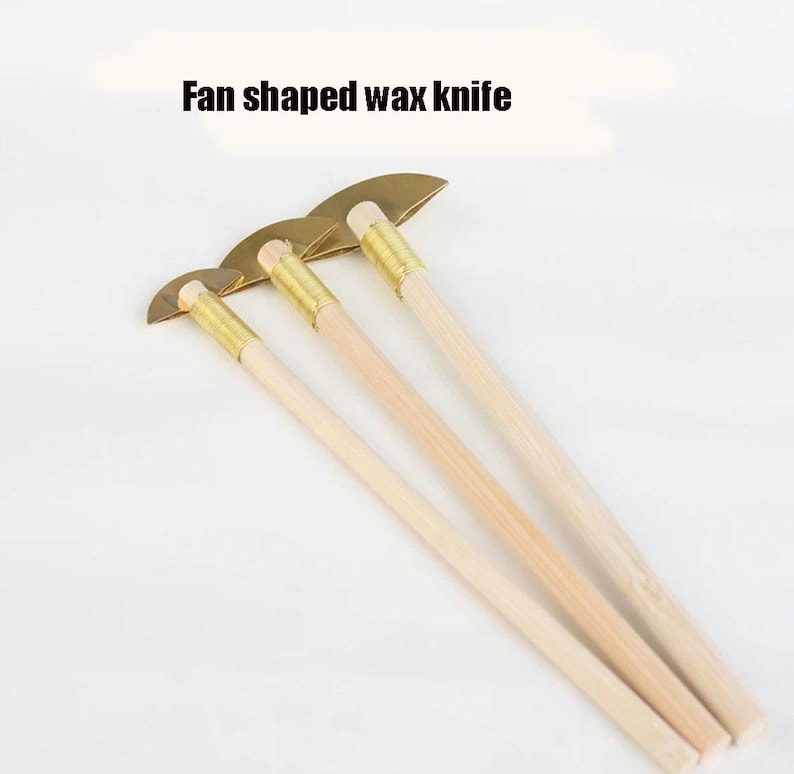 May include: Fan-shaped wax knives with wooden handles and brass-coloured blades. The text "Fan shaped wax knife" is displayed above the knives. The tools are designed for wax carving and sculpting.
