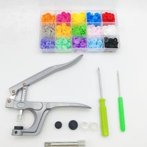 May include: A silver metal snap fastener plier tool, a clear plastic storage box containing fifteen compartments of colorful plastic snap buttons, two awls with green handles, and a small metal rod component for sewing and crafting projects.