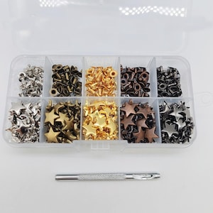 Peut inclure: Une boîte de rangement en plastique transparent à dix compartiments contenant des rivets métalliques et des clous en forme d'étoile aux finitions argentée, dorée, bronze et noire. Un outil de pose métallique est placé sous la boîte.