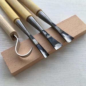 May include: A collection of wood carving tools featuring wooden handles and metal blades, including a hook knife and gouges. The tools are displayed on a small, rectangular block of light-coloured wood.