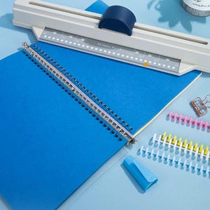 May include: A white 30-hole paper punch tool with a dark blue sliding handle positioned above a blue notebook with a metal spiral binding. Nearby are several plastic binding spines in pink, yellow, and white, plus a small blue plastic tool.
