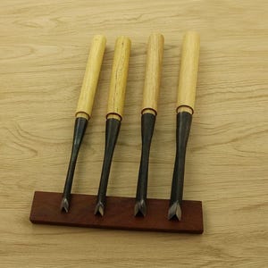 Puede incluir: Cuatro formones de madera con mangos de madera de color claro y hojas de metal oscuro se exhiben sobre una base de madera marrón oscuro. Los formones están en ángulo, mostrando sus bordes afilados y biselados. El fondo es una superficie de madera clara.