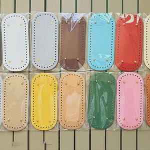 May include: A collection of fourteen rectangular bag bases with rounded ends, each featuring pre-punched holes along the perimeter and four small metal feet. The bases are available in various solid colors including black, white, gray, brown, blue, red, yellow, green, and pink.