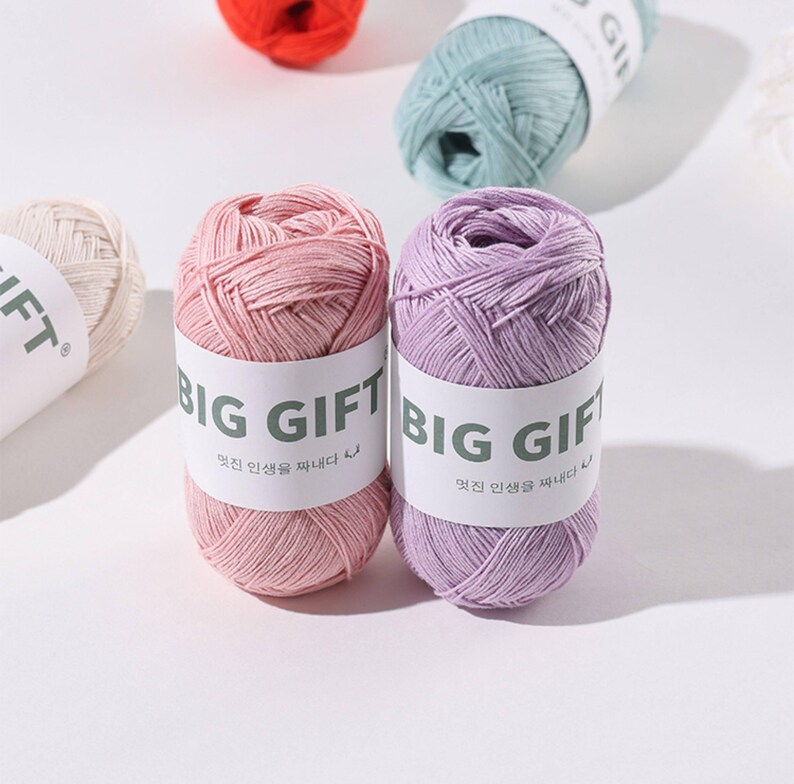 May include: Two skeins of yarn in pink and light purple, each wrapped in a white paper band labeled BIG GIFT with Korean text. Additional skeins in cream, light blue, and red are partially visible in the background, all featuring the same label.