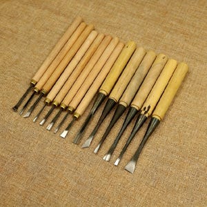 May include: A set of wood carving chisels with wooden handles and metal blades. The chisels are arranged diagonally on a tan fabric surface. The blades have various shapes for different carving needs.
