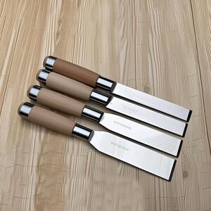 Peut inclure: Quatre spatules en acier inoxydable avec des manches en bois et des accents argentés sont disposées en diagonale sur une surface en bois. Les spatules ont une forme de lame rectangulaire et sont gravées de texte. Les manches sont marron clair et cylindriques.