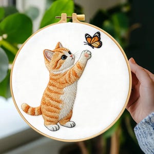 May include: Embroidered hoop art featuring a ginger tabby kitten reaching for a butterfly. The kitten has blue eyes and is rendered in detailed stitching. The butterfly is orange and black. The hoop is made of wood and the background is white.