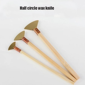 May include: Three half-circle wax knives with wooden handles and brass blades. The text "Half circle wax knife" is printed above the knives. The blades are attached to the handles with copper-coloured metal. The handles are made of light-coloured wood.
