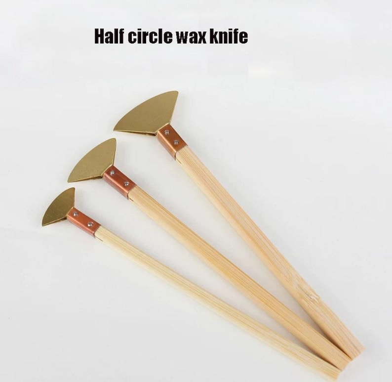 May include: Three half-circle wax knives with wooden handles and brass blades. The text "Half circle wax knife" is printed above the knives. The blades are attached to the handles with copper-coloured metal. The handles are made of light-coloured wood.