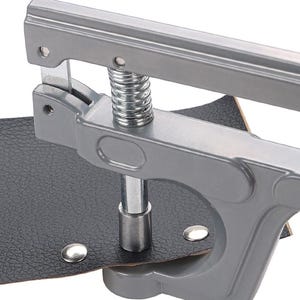 May include: A gray metal rivet setter tool with a spring mechanism, setting a silver rivet on black leather. The tool is designed for applying rivets to leather or similar materials, commonly used in crafting and repair.