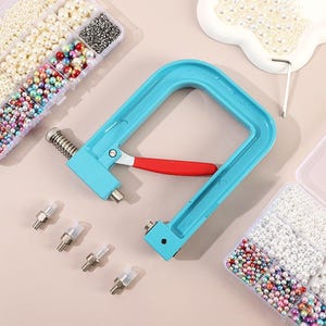 Puede incluir: Una herramienta manual azul claro con un mango rojo, diseñada para aplicar remaches. La imagen también muestra varios recipientes de plástico transparente llenos de perlas de varios tamaños y colores, junto con varias piezas de remache.