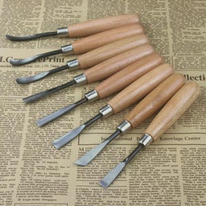 May include: A set of wood carving tools with metal blades and wooden handles. The tools are arranged on a newspaper background, showcasing their sharp edges and craftsmanship. Ideal for woodworking and detailed carving projects.