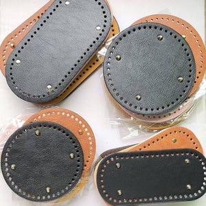 Op de afbeelding: Een verzameling ovale en ronde tassenbodems van gestructureerd zwart en bruin kunstleer. Elk stuk is voorzien van voorgeponste gaten langs de rand om te naaien en vier kleine metalen noppen aan de onderkant voor extra stabiliteit.