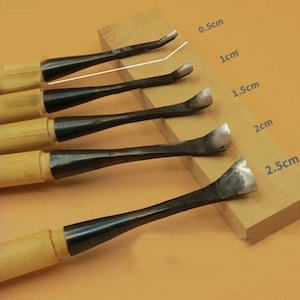May include: Five wood carving gouges with wooden handles and black metal blades. The blades range in size, from 0.5cm to 2.5cm. The tools are displayed on a wooden block, highlighting their varied shapes and sizes for detailed carving.