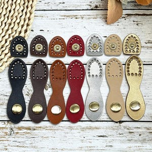 May include: A collection of leather-like tabs in black, brown, tan, red, silver, beige, and gold. The top row features small, rounded tabs with a central snap, while the bottom row shows elongated, hourglass-shaped tabs with a metallic snap button.
