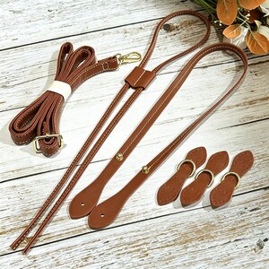 Peut inclure: Un ensemble d'accessoires de sac marron comprenant une longue bandoulière réglable avec des ferrures en métal doré, une sangle enroulée séparée et trois petites pattes en cuir marron avec des anneaux en D dorés pour sac à main.