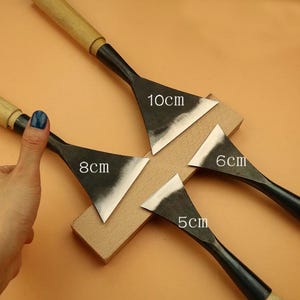 May include: Four metal woodworking chisels with wooden handles. The blades are triangular and range in size from 5cm to 10cm. The chisels are displayed on a light brown wooden block.