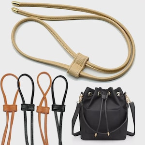 May include: A black bucket bag with a drawstring closure and a set of four adjustable bag straps in beige, tan, and black. Each strap features a sliding loop mechanism and gold-toned metal end caps for a secure and functional design.