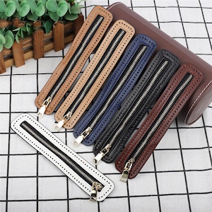 May include: A collection of six rectangular leather zipper pulls in white, tan, light brown, blue, black, and dark brown. Each piece features a central metal zipper with a gold-tone pull tab and pre-punched stitching holes along the edges.