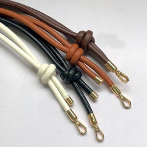 May include: Close-up of several leather bag straps in various colors: white, black, brown, and tan. Each strap features a knotted design and gold-colored metal hardware, including clasps and end caps. The straps are arranged diagonally.