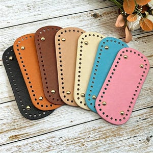 May include: A collection of seven rectangular faux leather bag bases in black, orange, brown, tan, cream, turquoise, and pink. Each base features rounded corners, a textured surface, pre-punched stitching holes along the edges, and four gold metal feet.
