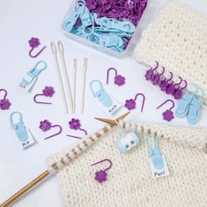 Può includere: Una collezione di strumenti e accessori per lavorare a maglia, tra cui segnapunti, misuratori di aghi e porta punti. I segnapunti sono viola e a forma di fiore, e i porta punti sono azzurri. I ferri da maglia sono in legno e il filato è bianco.