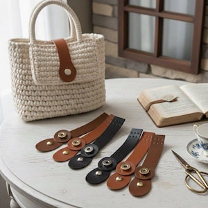 May include: A cream-colored crocheted handbag with two top handles and a brown leather-like strap closure featuring a silver-tone snap button. Five additional leather-like straps in brown and black with snap buttons are arranged on the table.
