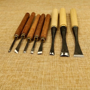 Puede incluir: Un juego de cinceles para tallar madera con mangos de madera y hojas de metal. Los cinceles tienen varias formas de hoja, incluyendo anguladas, planas y curvas. Los mangos son de color marrón oscuro y las hojas son plateadas y negras.