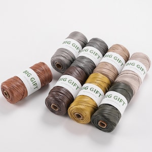May include: A collection of nine cylindrical spools of raffia ribbon in various earth tones including brown, gray, tan, and yellow. Each spool is wrapped with a white paper band featuring the text BIG GIFT in bold green capital letters.