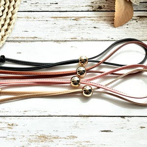 May include: Four thin, smooth cord straps in black, brown, light pink, and beige. Each strap features a polished gold-tone spherical bead that slides along the cord, with knotted ends to secure the loop, creating a simple adjustable design.