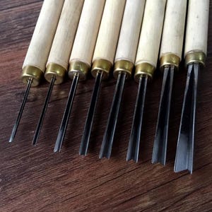 May include: A set of wood carving tools with wooden handles and metal blades. The tools have brass ferrules and are arranged in a row, showcasing various blade shapes for detailed woodworking projects.