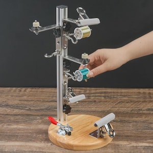 Puede incluir: Un organizador de hilo de metal plateado con base de madera. El organizador tiene múltiples brazos que sostienen carretes de hilo en varios colores, incluyendo verde y dorado. Se muestra una mano interactuando con los carretes de hilo. La base tiene una abrazadera y un clip blanco.