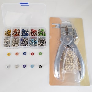 Puede incluir: Una herramienta perforadora de ojales de metal en un blíster con una bolsa de ojales dorados, junto a una caja de almacenamiento de plástico transparente con diez compartimentos llenos de ojales metálicos de colores amarillo, naranja, azul, rojo, verde y plateado.