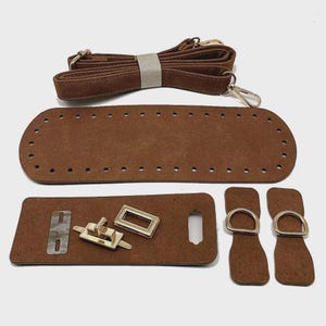 May include: A brown bag making kit featuring a long adjustable strap, a large rectangular base with pre-punched holes, a smaller rectangular flap with a metal turn lock, and two side tabs with attached metal D-rings for assembly.