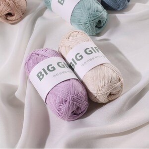 May include: Two skeins of yarn, one in light purple and one in cream, each wrapped in a white paper band labeled BIG GIFT with Korean text. Additional skeins in light blue and tan are visible in the background, all featuring the same label.
