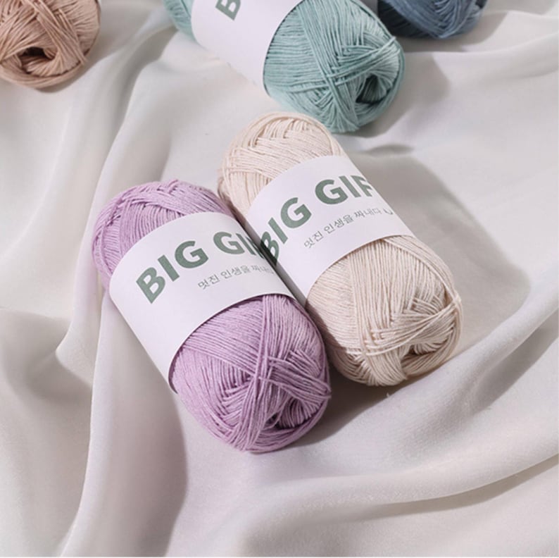 May include: Two skeins of yarn, one in light purple and one in cream, each wrapped in a white paper band labeled BIG GIFT with Korean text. Additional skeins in light blue and tan are visible in the background, all featuring the same label.