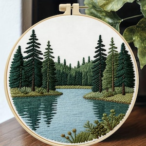 May include: Round embroidery hoop featuring a serene landscape. The design depicts a tranquil lake surrounded by lush green pine trees and foliage. The water reflects the trees, creating a mirrored effect. The embroidery is set against a white background.