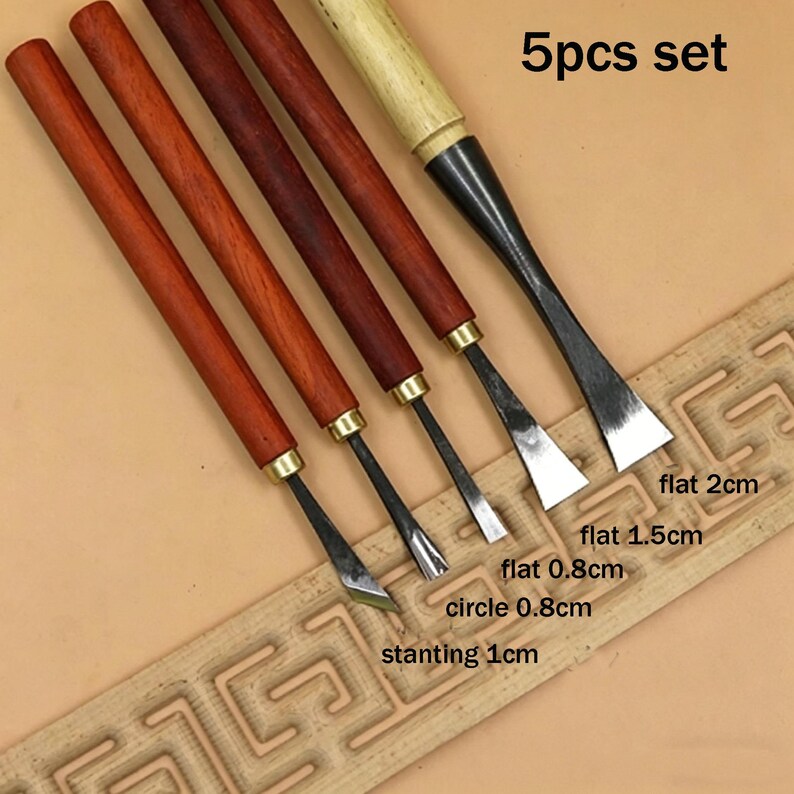 Op de afbeelding: Een set van vijf houtsnijgereedschappen. De gereedschappen hebben houten handvatten en metalen bladen. De set bevat platte, cirkelvormige en schuine beitels, met bladbreedtes van 0,8 cm tot 2 cm. De tekst "5pcs set" is zichtbaar.