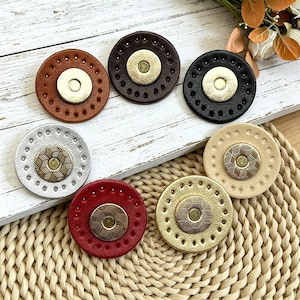 May include: A collection of seven circular magnetic fasteners in various colours including brown, dark brown, black, silver, red, gold, and beige. Each fastener features a central metallic button with a geometric pattern and a perforated border.