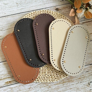 May include: Five oval-shaped bag bases in various colors: tan, black, brown, cream, and gray. Each base has a series of small holes around the edge for stitching and is secured with brass-colored rivets. The bases are arranged on a woven mat.