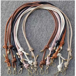 Könnte beinhalten: Eine Auswahl an Leder-Taschenriemen in verschiedenen Farben, darunter Braun, Schwarz, Weiß und Rosa. Jeder Riemen hat einen Knoten und goldfarbene Metallverschlüsse. Sie liegen auf einem grauen Hintergrund.