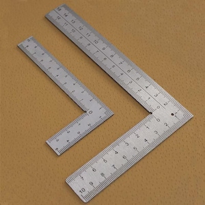 Puede incluir: Tres escuadras de metal plateado con marcas de medición grabadas. La escuadra más grande mide 38 cm de largo, la mediana 25 cm y la más pequeña 10 cm. Las escuadras están dispuestas sobre una superficie marrón.