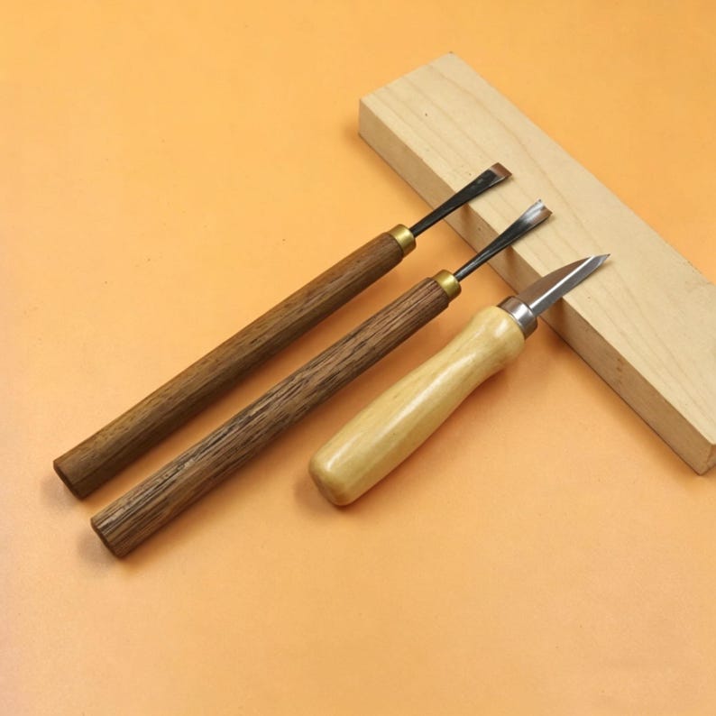 Op de afbeelding: Drie houtsnijgereedschappen met metalen messen en houten handvatten. Twee gereedschappen hebben donkerbruine handvatten en &eacute;&eacute;n heeft een lichtbruin handvat. Een lichtgekleurd houten blok rust op de achtergrond tegen een oranje achtergrond.