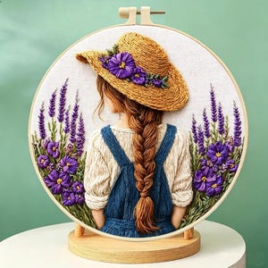 Puede incluir: Obra de arte bordada en un aro de madera que representa a una persona con un sombrero de paja adornado con flores moradas, de espaldas. La persona tiene una larga trenza y lleva un peto azul. Flores de lavanda y moradas enmarcan la figura, creando una escena serena.