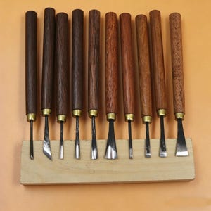 Op de afbeelding: Een set van tien houtsnijgereedschappen met donkerbruine houten handvatten en messing hulzen. De bladen hebben verschillende vormen en maten en worden gepresenteerd op een lichtgekleurd houten blok. De gereedschappen zijn in een rij gerangschikt.