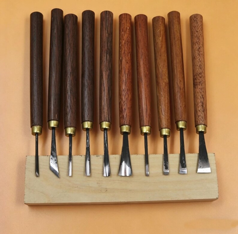 Op de afbeelding: Een set van tien houtsnijgereedschappen met donkerbruine houten handvatten en messing hulzen. De bladen hebben verschillende vormen en maten en worden gepresenteerd op een lichtgekleurd houten blok. De gereedschappen zijn in een rij gerangschikt.
