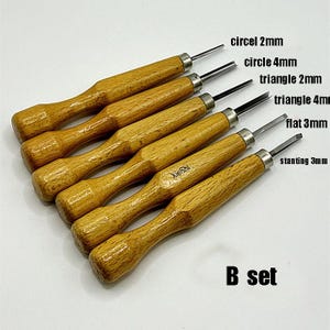 Puede incluir: Un juego de seis herramientas para tallar madera con mangos de madera y cuchillas de metal. Las cuchillas están etiquetadas con sus formas y tamaños, incluyendo círculo 4mm, triángulo 2mm y plano 3mm. El texto "B set" está en la parte inferior.