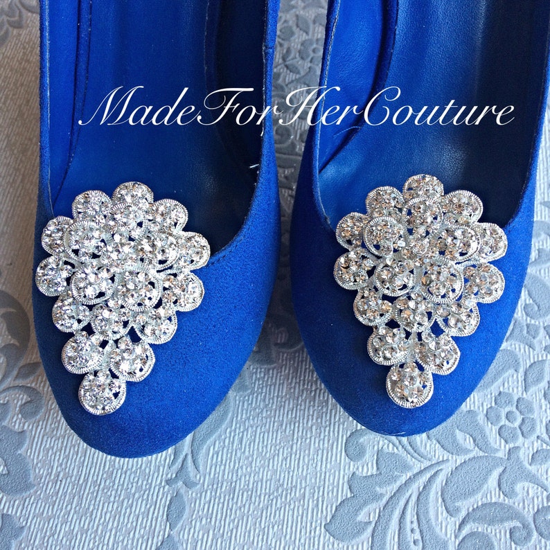 K&ouml;nnte beinhalten: Ein Paar blaue Wildleder-High-Heel-Schuhe mit silbernen Strasssteinen an den Zehen. Die Verzierungen sind in Form einer Blumenansammlung gestaltet.