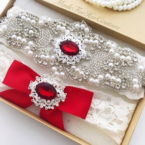 May include: A white lace garter with a red satin bow and two silver and rhinestone embellishments with red gems. The garter is in a box with the text "Made For Her Couture".